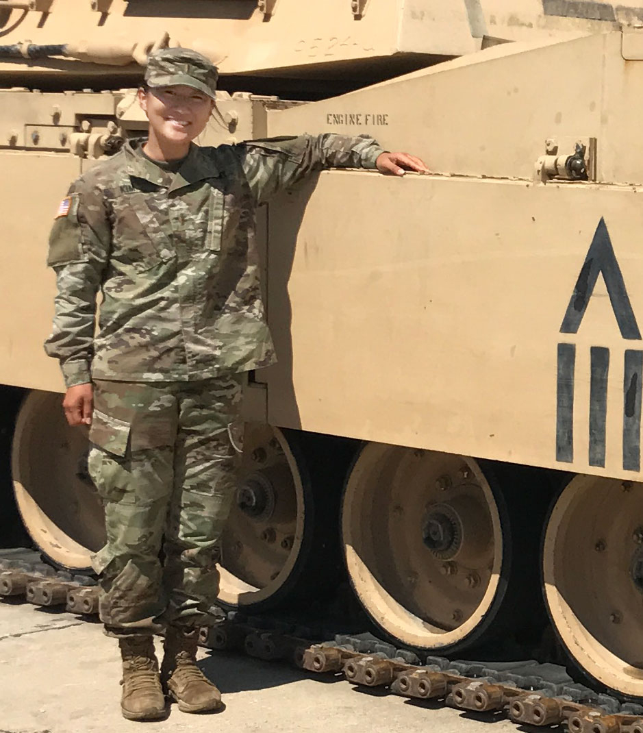 Esther Kim, a 2016 Fordham graduate and one of the first women allowed into combat roles in the U.S. military, is shown dressed in Army fatigues and standing next to a tank. 