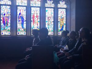 Students sit in pews in the Blue Chapel at the Rose Hill campus, with their eyes closed and their palms facing up; behind them are stained glass windows.