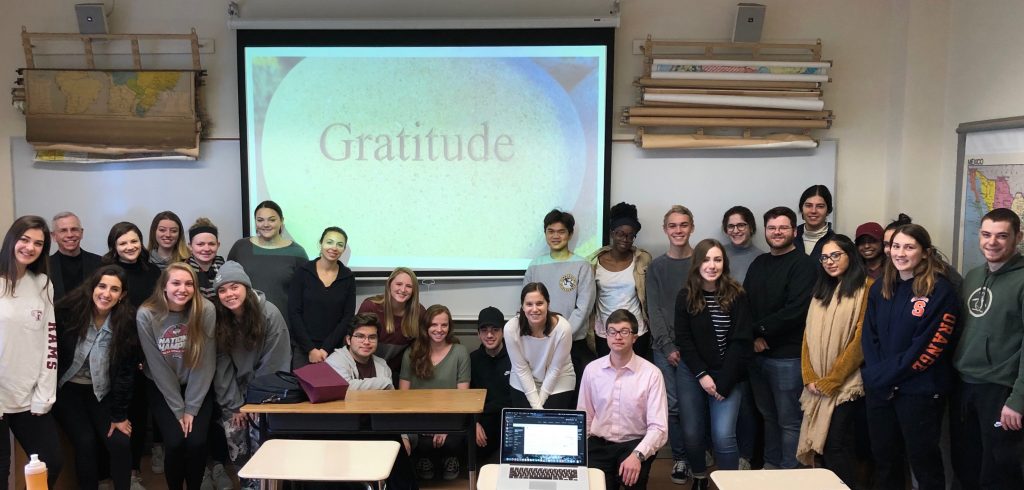 Father Marcotte's class poses for a group picture at the front of the classroom. They are standing below a projector screen that has a picture of a stone with the engraved word "Gratitude."