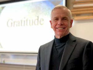 Father Marcotte smiles next to an image that says the word "Gratitude."