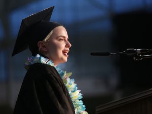 A girl with short blonde hair and a black graduation gown speaks in front of a microphone