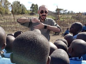 A man wearing sunglasses speaks and gestures with his hands in front of a crowd of Kenyan children