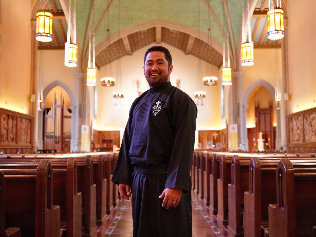A man wearing black priest robes stands in the middle of the University Church, between the church pews