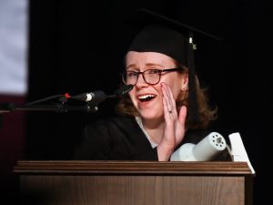 A girl wearing glasses speaks at a podium while holding a hand to the side of her face