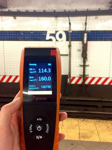 A hand holding an orange device in front of a subway station wall that says "59"