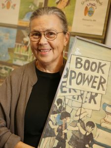 A woman wearing glasses smiles and holds a photo that says "BOOK POWER."