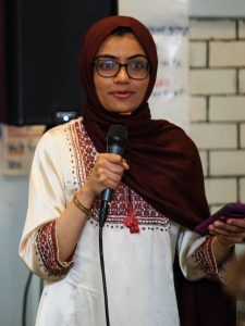 A woman wearing a hijab and glasses speaks into a microphone.