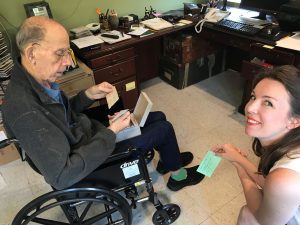 A man sitting in a wheelchair, holding a box of index cards, and a smiling woman beside him