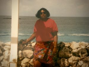 A woman wearing sunglasses, a red shirt, and a floral skirt stands in front of the ocean.