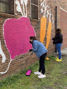 A woman wearing a jean jacket and black pants paints a purple beet on a brick wall.