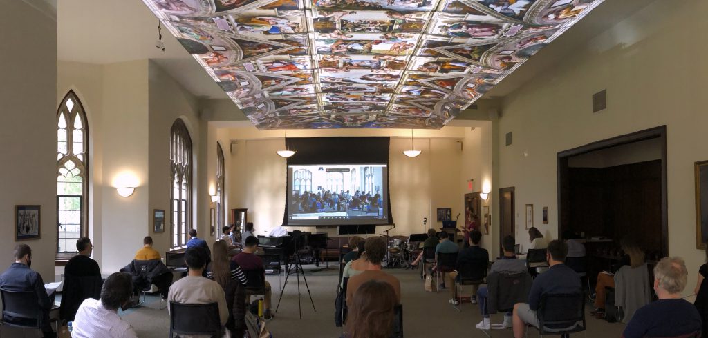 People sit in chairs spaced six feet apart in a large room with a painting on the ceiling.