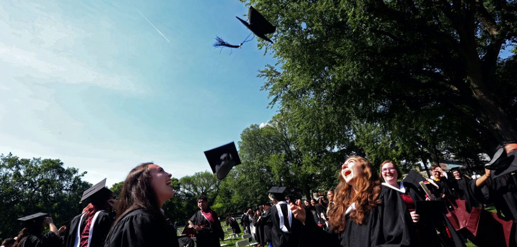Students wearing black graduation gowns watch a black graduation cap being tossed into the sky.