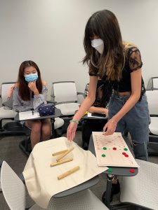 Two students wearing masks look at a pile of sticks (a board game) on a classroom desk.