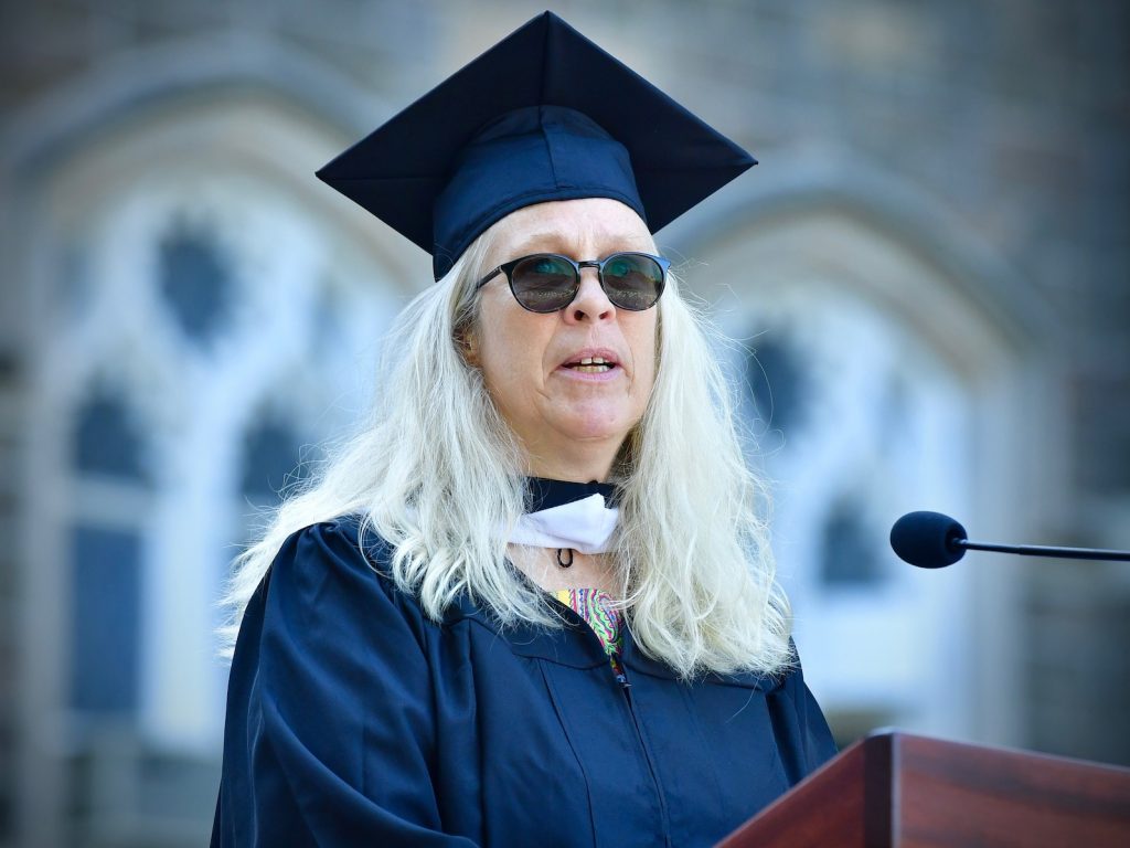 A woman with white blonde hair, sunglasses, and a graduation gown speaks at a wooden podium.