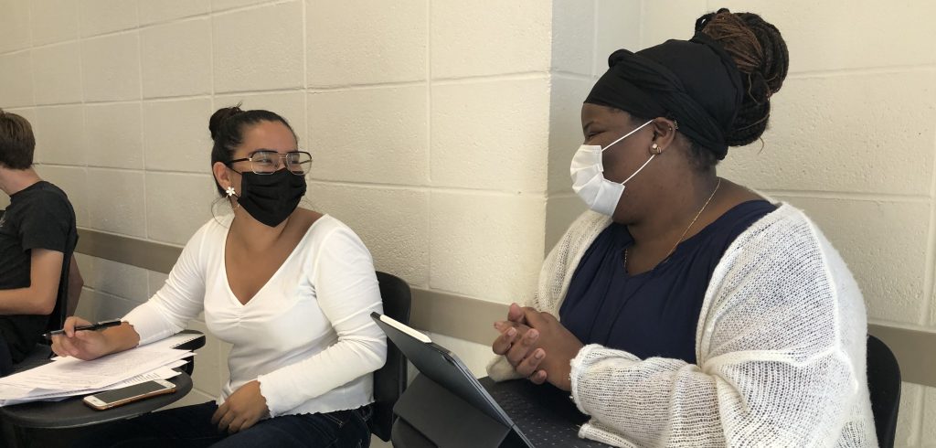 Two students wearing masks smile at each other in a classroom.